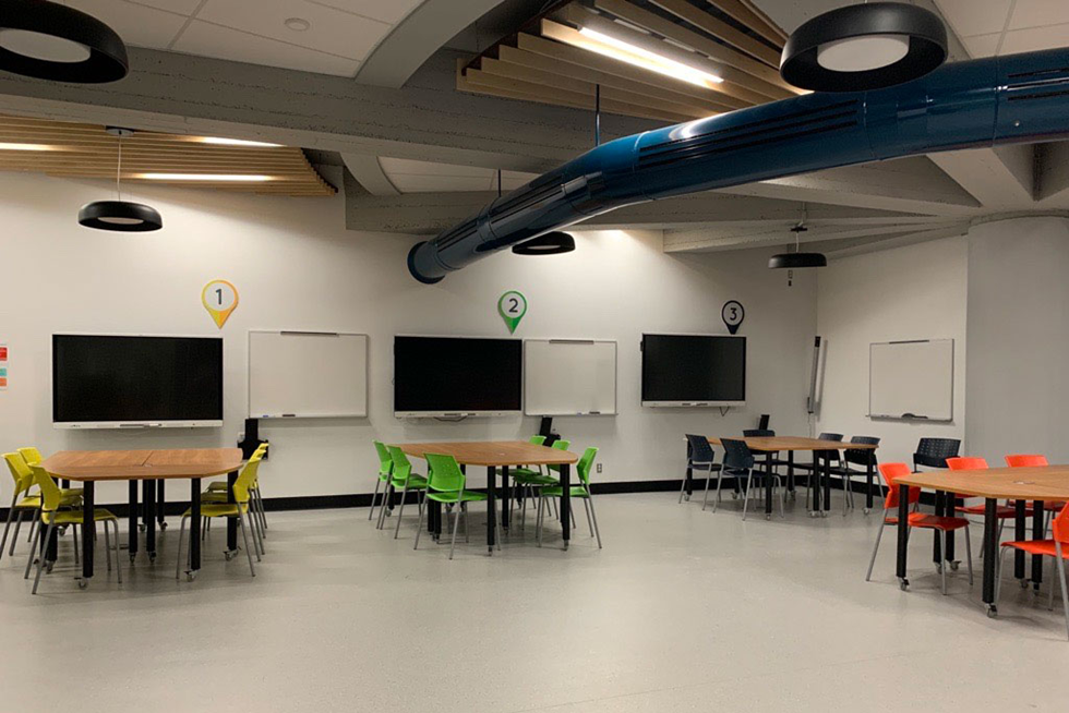 La 1re CLAAC du Cégep de Jonquière. Dans un grand local de classe, on voit 4 ilots de tables modulables entourés chacun de 6 chaises, dont les couleurs changent d’un ilot à l’autre. Au mur, au-dessus de chaque ilot, un numéro dans une bulle de la même couleur que les chaises de l’ilot correspondant. Sous chaque numéro, il y a un téléviseur intelligent et un tableau blanc.